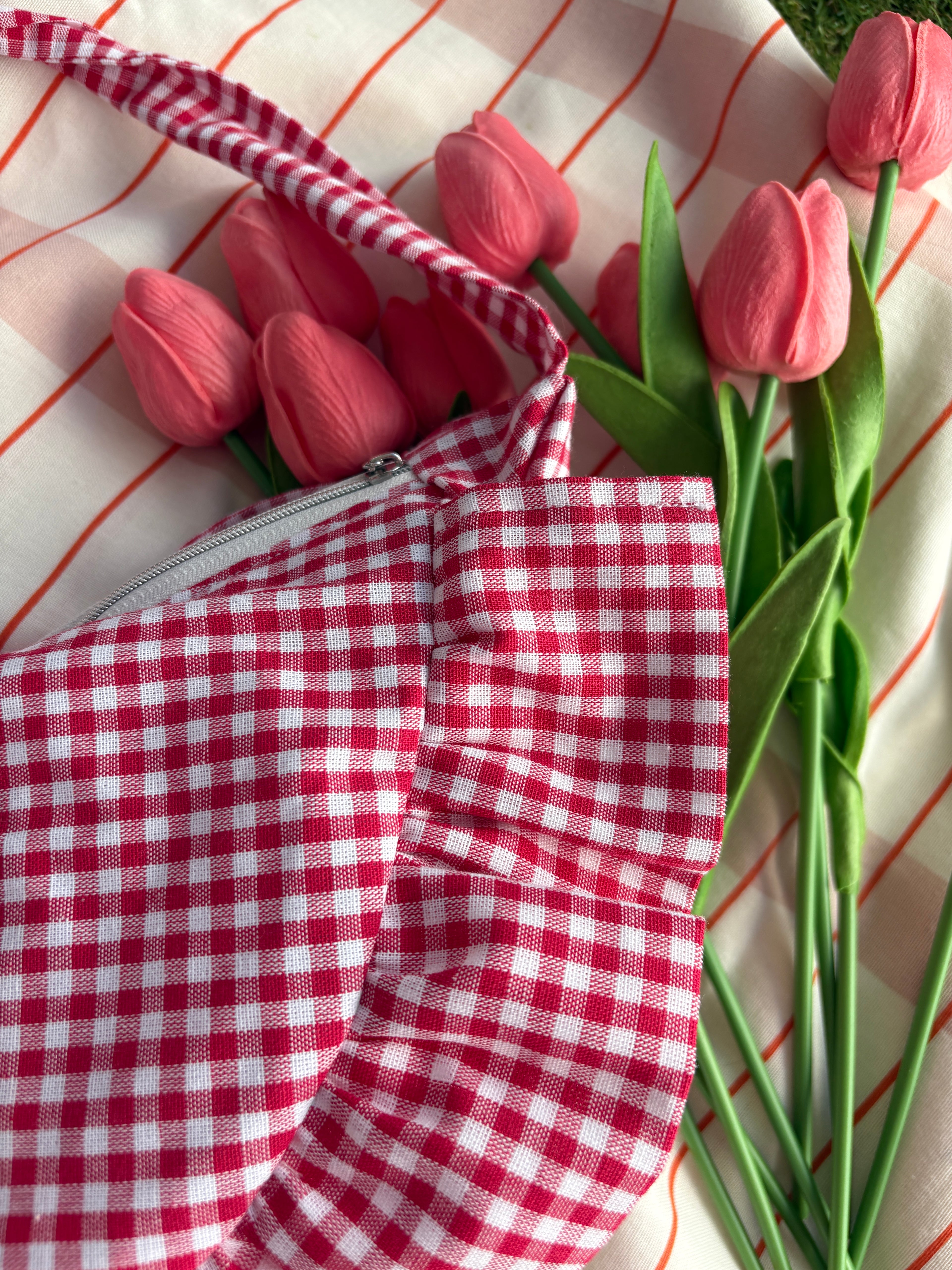 Plan rapproché sur les détails du sac à main à volants en forme de demi-lune/croissant, fait main, coloris Fraise, collection Printemps, sur une nappe de pique-nique, avec des tulipes autour du sac de la marque SIOU