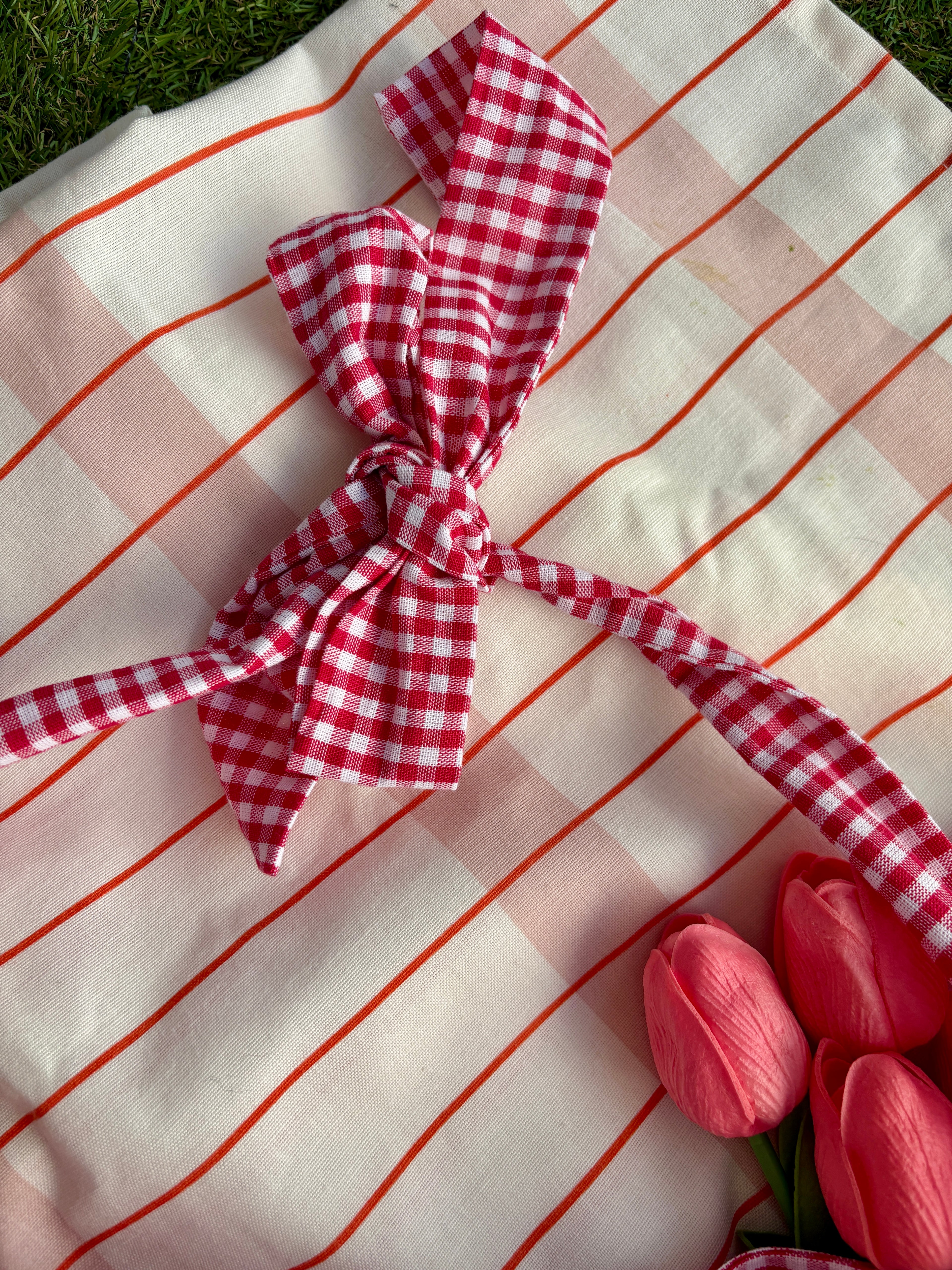 Zoom sur le nœud du sac à main à volants artisanal en forme de demi-lune/croissant, coloris Fraise, collection Printemps, sur une nappe de pique-nique, sur de l'herbe avec des tulipes autour du sac en tissu de la marque SIOU
