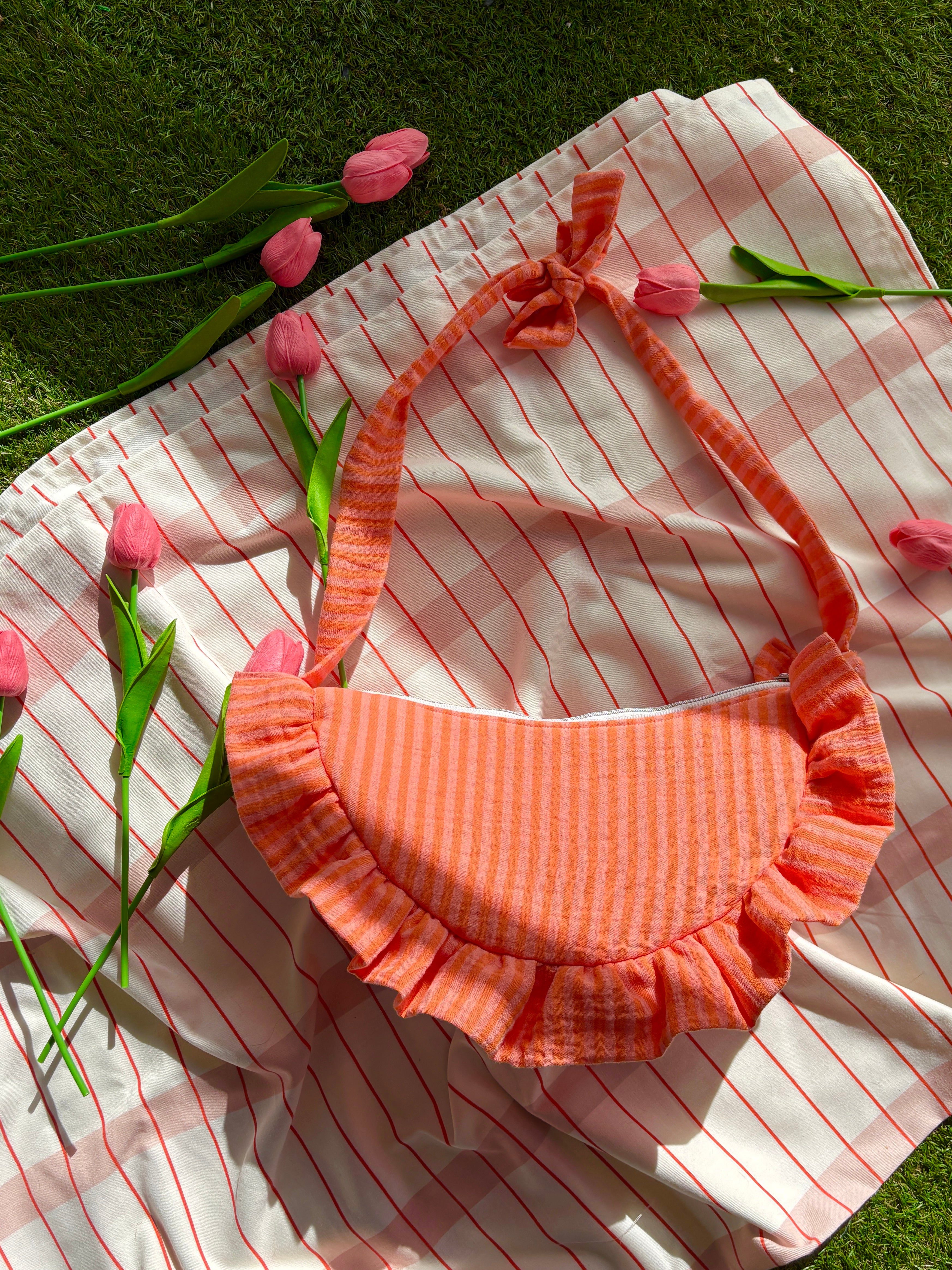 Sac à main en tissu gaze de coton à volants en forme de croissant, cousu main, coloris rayures orange et rose Pêche-Abricot, collection Printemps, sur une nappe de pique-nique, sur de l'herbe avec des tulipes autour du sac de la marque SIOU