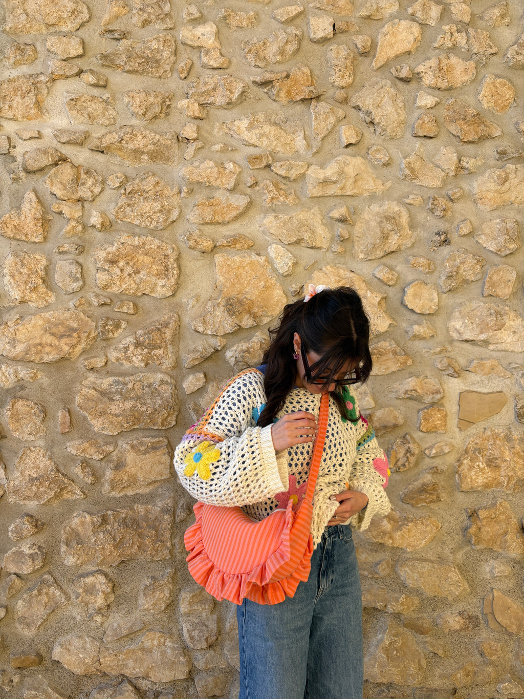 Sac à main SIOU en tissu gaze de coton à volants en forme de demi-lune, fait main, coloris rayures orange et rose Pêche-Abricot, collection Printemps, porté en bandoulière par une jeune femme habillée par un pull à fleurs colorées en mailles et un jean debout devant un mur en pierre provençal.