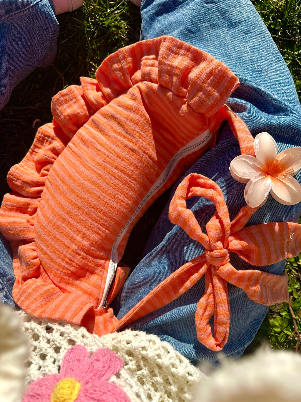 Sac à main SIOU en tissu gaze de coton à volants en forme de croissant, artisanal, coloris rayures orange et rose Pêche-Abricot, collection Printemps, posé entre les jambes d'une fille vêtue d'un pull en mailles coloré et une paire de jeans bleu. La anse du sac est décorée d'une pince à cheveux orange et blanc en forme de fleur.