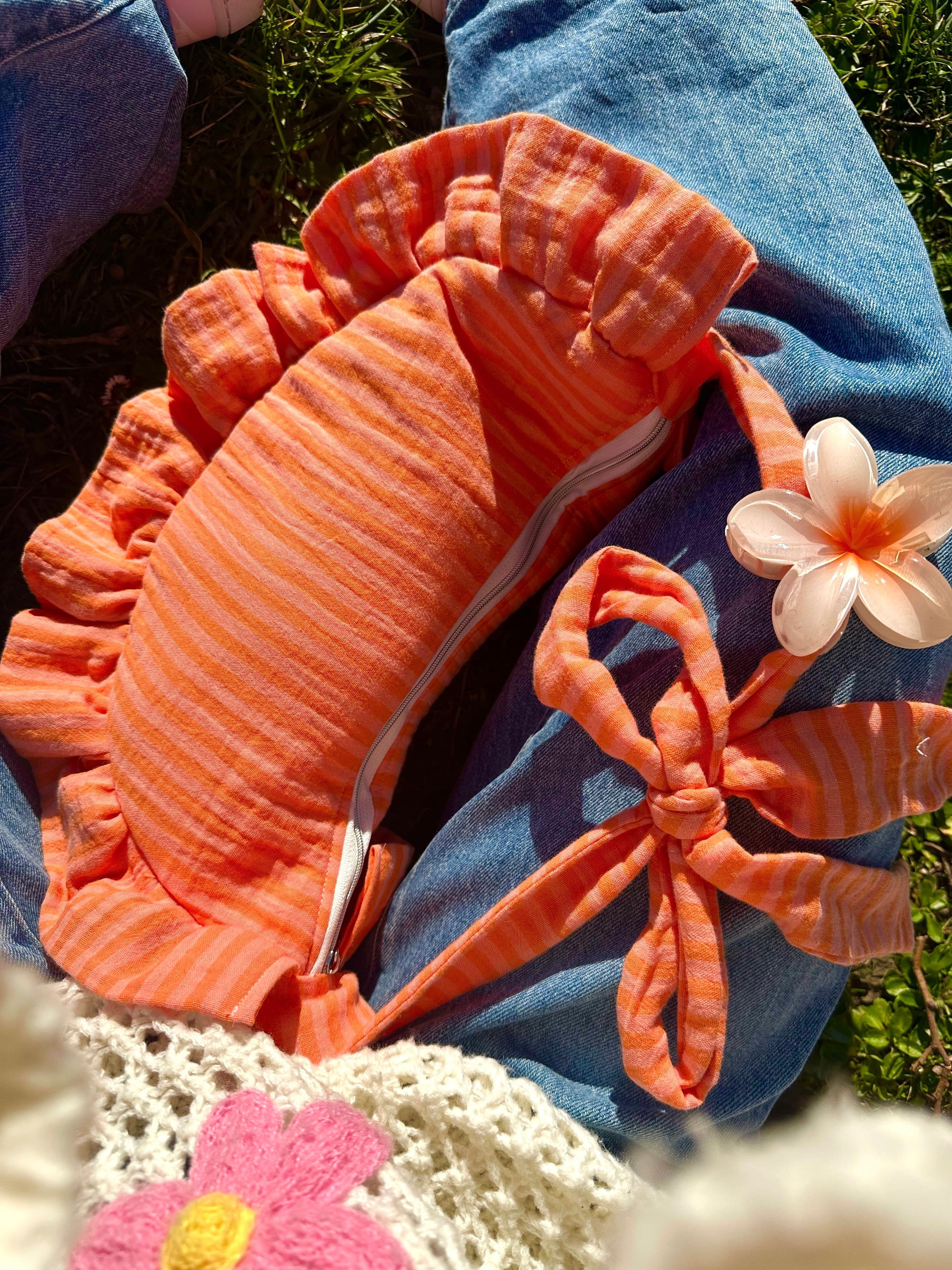 Sac à main SIOU en tissu gaze de coton à volants en forme de croissant, artisanal, coloris rayures orange et rose Pêche-Abricot, collection Printemps, posé entre les jambes d'une fille vêtue d'un pull en mailles coloré et une paire de jeans bleu. La anse du sac est décorée d'une pince à cheveux orange et blanc en forme de fleur.