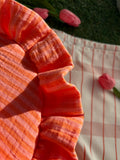 Zoom sur les détails de couture du sac à main en tissu gaze de coton à froufrous en forme de demi-lune, cousu main, coloris rayures orange et rose Pêche-Abricot, collection Printemps, sur une nappe de pique-nique, sur de l'herbe avec des tulipes autour du sac de la marque SIOU