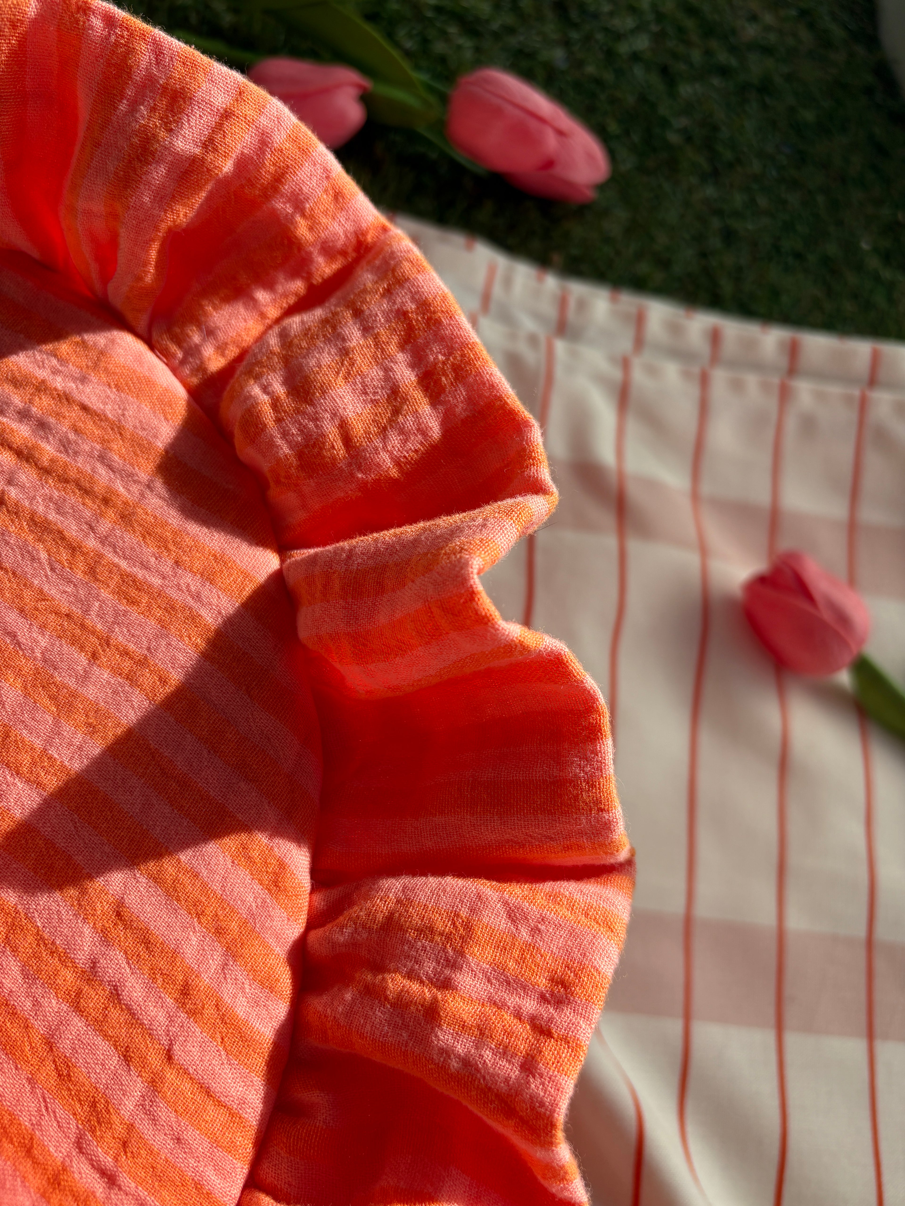 Zoom sur les détails de couture du sac à main en tissu gaze de coton à froufrous en forme de demi-lune, cousu main, coloris rayures orange et rose Pêche-Abricot, collection Printemps, sur une nappe de pique-nique, sur de l'herbe avec des tulipes autour du sac de la marque SIOU