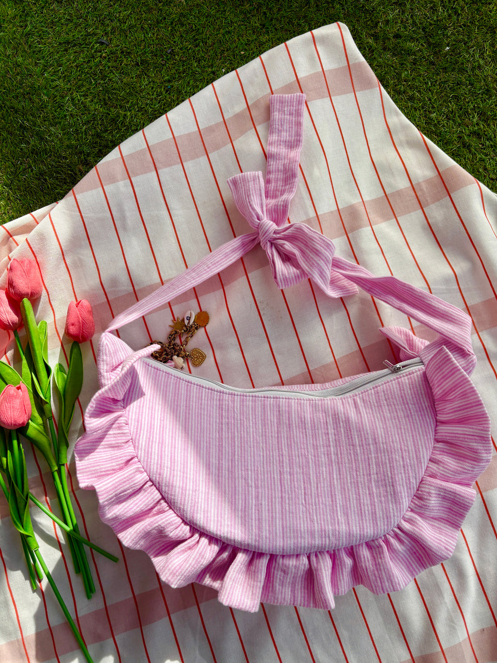 Sac à main en tissu gaze de coton à volants en forme de croissant, cousu main, coloris rayures rose et blanc Tulipe, collection Printemps, sur une nappe de pique-nique, sur de l'herbe avec des tulipes autour du sac de la marque SIOU