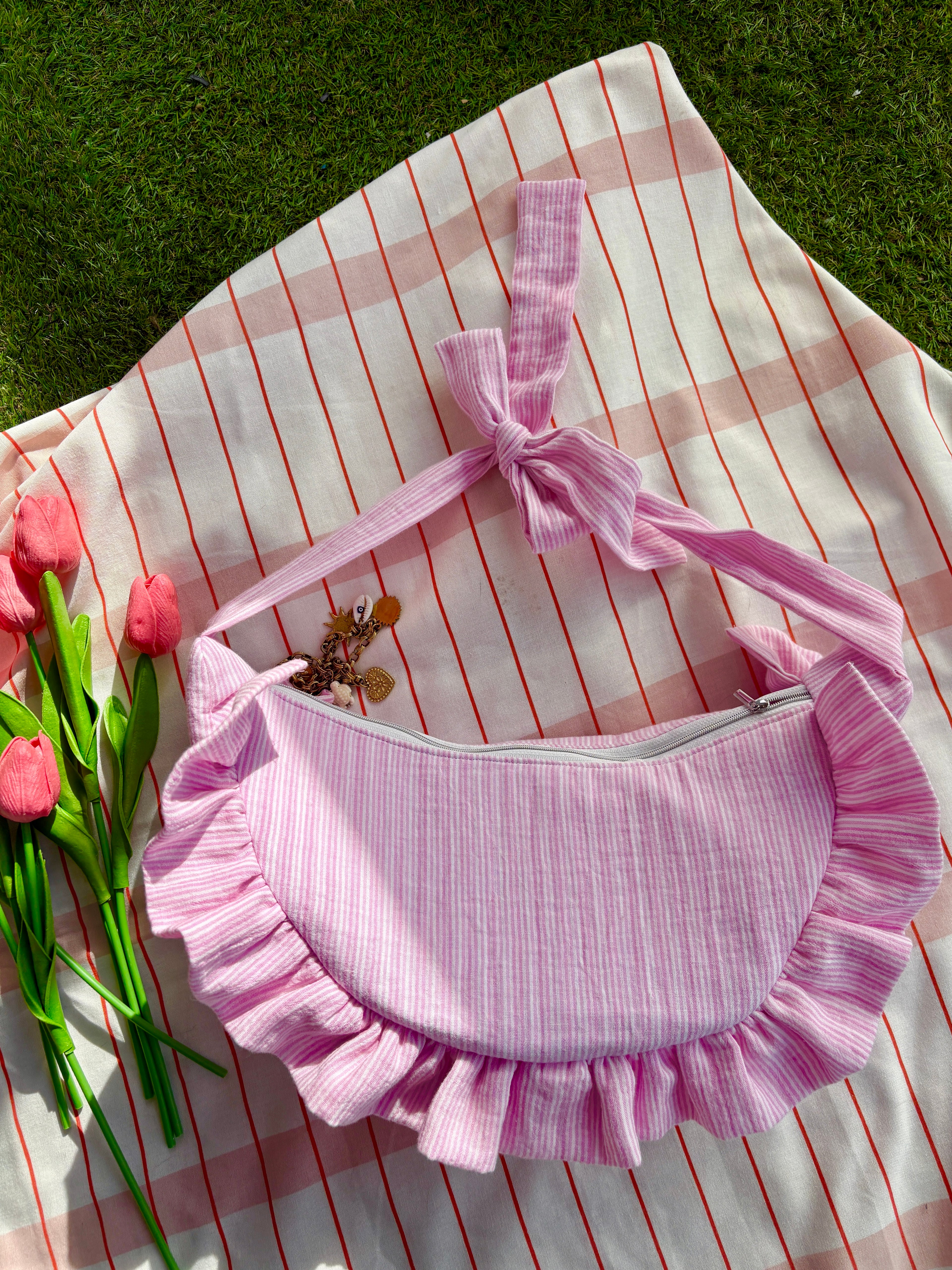 Sac à main en tissu gaze de coton à volants en forme de croissant, cousu main, coloris rayures rose et blanc Tulipe, collection Printemps, sur une nappe de pique-nique, sur de l'herbe avec des tulipes autour du sac de la marque SIOU
