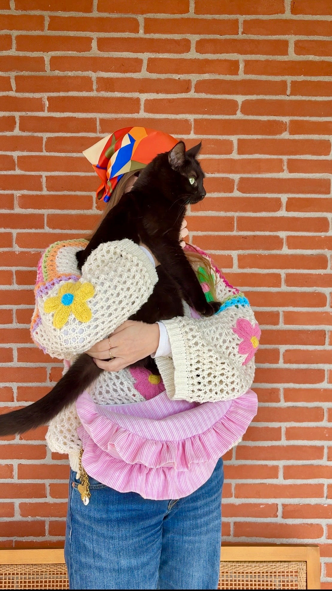 Sac à main en tissu gaze de coton à volants en forme de demi-lune, fait main, coloris rayures rose et blanc Tulipe, collection Printemps, porté en bandoulière par une femme portant une tenue colorée et un chat noir noir dans les bras devant un mur de briques rose de Toulouse