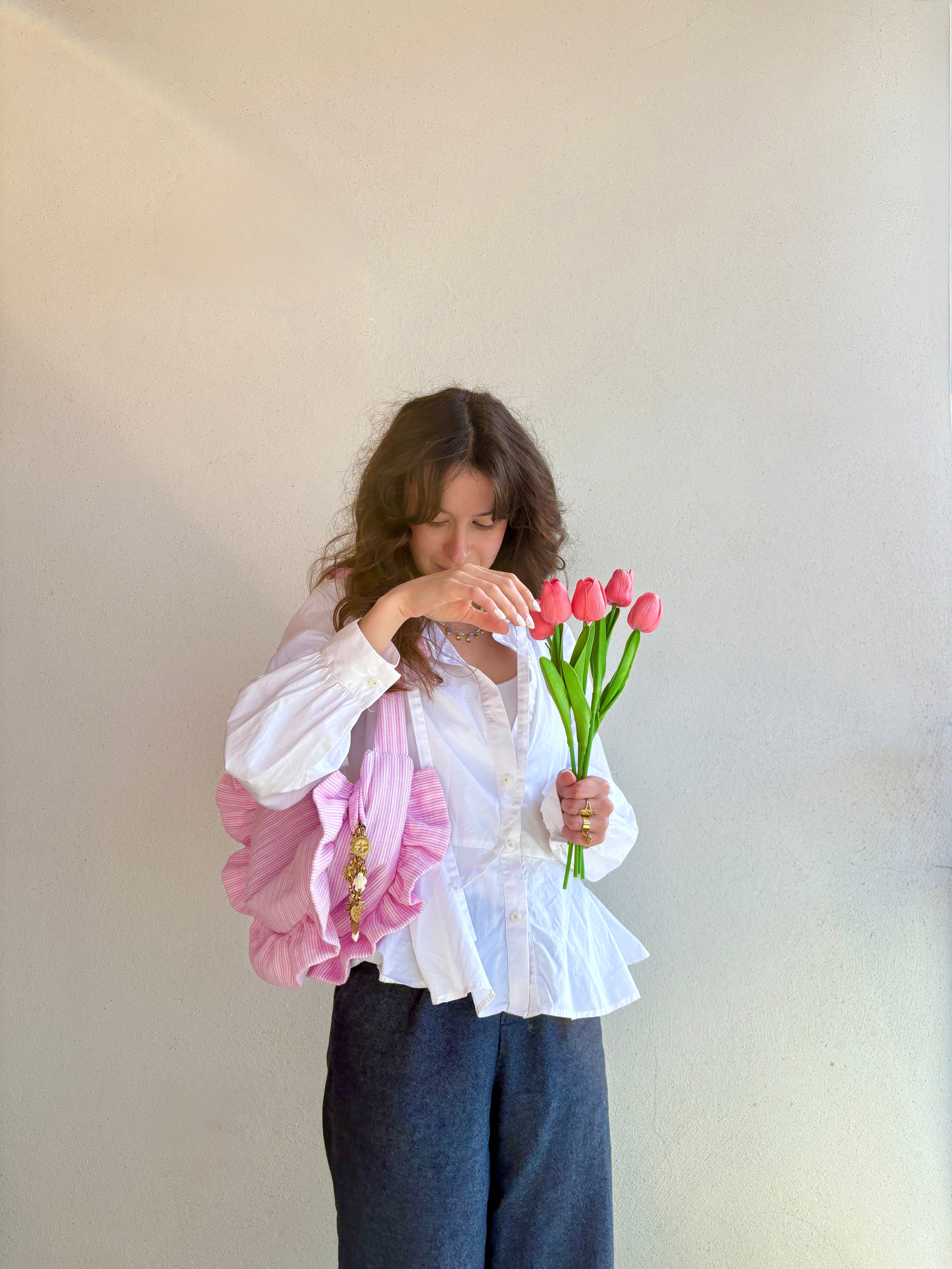 Sac à main en tissu gaze de coton à volants en forme de croissant, artisanal, coloris rayures rose et blanc Tulipe, collection Printemps, porté à l'épaule par une femme vêtue d'une chemise blanche et d'un jean avec un bouquet de tulipe roses dans la main